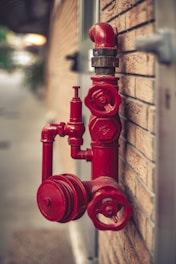 A red fire hydrant system is mounted on a brick wall. The hydrant has multiple valves and connectors, with one large valve in the foreground and a pipe extending upwards.