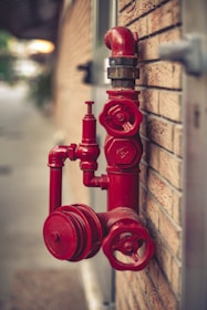 A red fire hydrant system is mounted on a brick wall. The hydrant has multiple valves and connectors, with one large valve in the foreground and a pipe extending upwards.