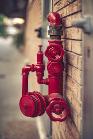 A red fire hydrant system is mounted on a brick wall. The hydrant has multiple valves and connectors, with one large valve in the foreground and a pipe extending upwards.