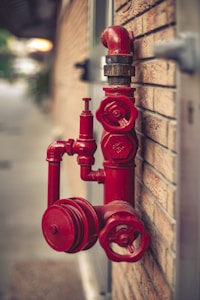 A red fire hydrant system is mounted on a brick wall. The hydrant has multiple valves and connectors, with one large valve in the foreground and a pipe extending upwards.