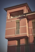 A modern residential building with a pink exterior. The structure features rectangular windows with white railings and a flat roof. The building is bathed in warm sunlight, casting soft shadows.