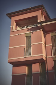 A modern residential building with a pink exterior. The structure features rectangular windows with white railings and a flat roof. The building is bathed in warm sunlight, casting soft shadows.