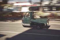 A small green utility vehicle with an open cargo bed is driving down a road. The background appears blurred, suggesting motion, with buildings and greenery indistinct in the distance.