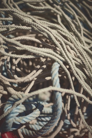 A tangle of ropes with varying textures and colors, primarily in shades of blue and beige. The ropes are intertwined in a complex and chaotic pattern, with some sections catching light and casting shadows.