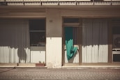 A building facade with two large windows covered by light curtains. A turquoise cloth is draped over an air conditioning unit next to a window. The building exterior is weathered, and there is a small rectangular planter with a few plants on the ground.