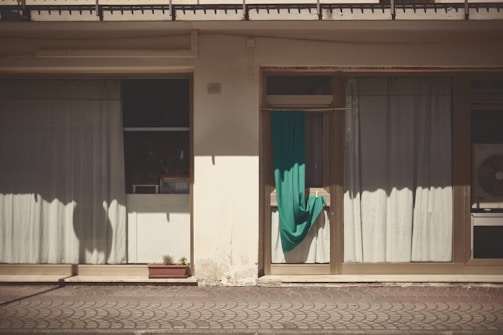 A building facade with two large windows covered by light curtains. A turquoise cloth is draped over an air conditioning unit next to a window. The building exterior is weathered, and there is a small rectangular planter with a few plants on the ground.