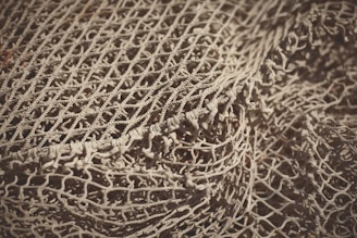 An intricate and detailed view of a loosely arranged fishing net, showcasing its intertwined patterns and textured knots. The net appears to be a light beige color with shadows accentuating its woven structure.