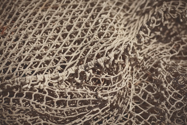 An intricate and detailed view of a loosely arranged fishing net, showcasing its intertwined patterns and textured knots. The net appears to be a light beige color with shadows accentuating its woven structure.
