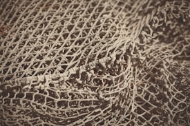 An intricate and detailed view of a loosely arranged fishing net, showcasing its intertwined patterns and textured knots. The net appears to be a light beige color with shadows accentuating its woven structure.