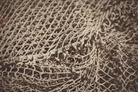 An intricate and detailed view of a loosely arranged fishing net, showcasing its intertwined patterns and textured knots. The net appears to be a light beige color with shadows accentuating its woven structure.