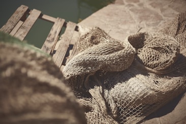 A rustic, aged wooden dock is situated beside a water body with a large, weathered fishing net draped over it. The textures of the wood and netting suggest a well-used and weather-beaten appearance, casting intricate shadows.