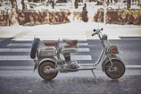 A vintage scooter with a side-mounted spare tire and brown leather seats is parked on a paved street. The scooter has a classic design with metallic components and a retro aesthetic. A striped pedestrian crossing is visible in the background, along with some brick structures and trees lining the street.