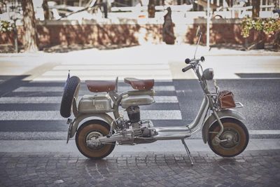 A vintage scooter with a side-mounted spare tire and brown leather seats is parked on a paved street. The scooter has a classic design with metallic components and a retro aesthetic. A striped pedestrian crossing is visible in the background, along with some brick structures and trees lining the street.