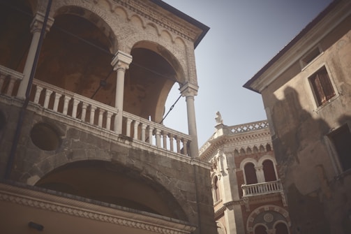 Architectural buildings featuring arched windows and intricate stonework create a blend of classical and Renaissance styles. A partly cloudy sky can be seen in the background, with sunlight casting shadows on the surfaces.