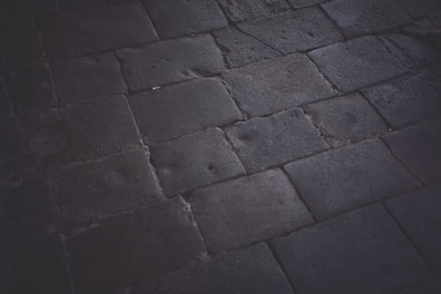 A dark, textured pavement composed of large rectangular stones. The stones are laid out in a simple geometric pattern, with visible wear and subtle marks on their surfaces indicating frequent use. The lighting is dim, casting soft shadows that highlight the texture of the stone material.