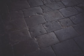 A dark, textured pavement composed of large rectangular stones. The stones are laid out in a simple geometric pattern, with visible wear and subtle marks on their surfaces indicating frequent use. The lighting is dim, casting soft shadows that highlight the texture of the stone material.