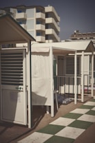 A row of small, beachside cabins with white walls and green roofs line a sandy area. The foreground features a checkered pathway made of white and green tiles, with a bright and sunny sky overhead. In the background, a tall residential building is visible.