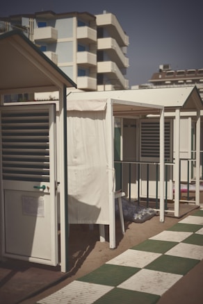 A row of small, beachside cabins with white walls and green roofs line a sandy area. The foreground features a checkered pathway made of white and green tiles, with a bright and sunny sky overhead. In the background, a tall residential building is visible.