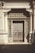 An ornate, intricately designed doorway with classical architectural elements. The heavy wooden door features square panels and metal accents, framed by tall columns with decorative capitals. Above the door is a decorative grille with elaborate patterns, and the structure is bathed in warm sunlight, casting shadows on the ground.