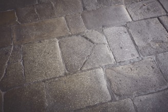 Weathered stone pavement with rectangular and irregular shapes, displaying varying textures and shades. Some stones show visible cracks and signs of wear, indicating age and use.