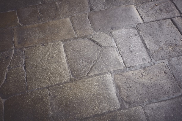 Weathered stone pavement with rectangular and irregular shapes, displaying varying textures and shades. Some stones show visible cracks and signs of wear, indicating age and use.
