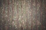 Old, weathered metal surface with vertical ridges, displaying a mix of green, brown, and rust colors. The surface shows signs of wear and peeling paint, creating a textured and rugged appearance.