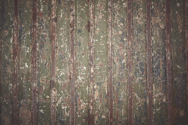 Old, weathered metal surface with vertical ridges, displaying a mix of green, brown, and rust colors. The surface shows signs of wear and peeling paint, creating a textured and rugged appearance.