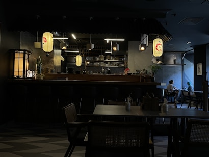 A dimly lit restaurant interior with an inviting atmosphere. Traditional hanging lanterns provide a warm glow, highlighting the bar area stocked with dishes and utensils. A chef or staff member in a red bandana can be seen behind the counter. Two people are seated at a table near the window, engaged in conversation.