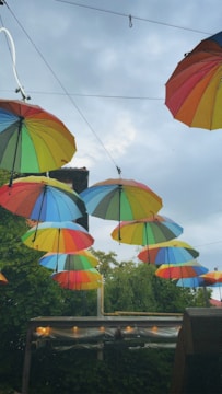 Multiple colorful umbrellas are hanging upside down in an outdoor setting. The umbrellas create a vibrant canopy against a backdrop of cloudy skies. Beneath the umbrellas, there are trees and some outdoor structures, adding a natural element to the scene.