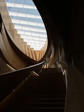A modern interior staircase with sleek wooden railings leads upwards towards a large, geometric skylight. The architecture features curved lines and natural light, emphasizing a sense of openness and innovation.