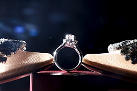 A sparkling diamond ring is the focal point, placed centrally between two pairs of high-heeled shoes with crystalline embellishments under a dramatic spotlight. The background is dark, enhancing the shine and elegance of the jewelry and shoes.