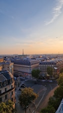 An aerial view of a cityscape featuring classic Parisian architecture with rows of uniform, multi-story buildings topped with mansard roofs. In the background, the Eiffel Tower stands prominently, set against a clear sky with a soft gradient of blue and orange hues from the setting sun. The foreground shows a circular intersection with streets branching out and surrounded by lush green trees, indicating a mix of urban and natural elements.