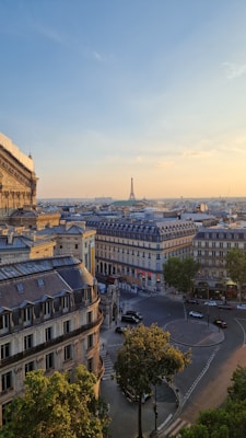 A scenic view of Paris during sunset, featuring elegant buildings with classic French architecture and the Eiffel Tower visible in the distance. The sky is clear with a warm glow, and the streets below show light traffic and a tranquil atmosphere.