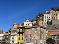 A series of colorful, traditional Mediterranean buildings are stacked along a hillside under a clear blue sky. The structures feature a mix of architectural styles, with pastel hues and rustic facades. Windows with shutters, tiled roofs, and satellite dishes are visible, adding character to the scene.