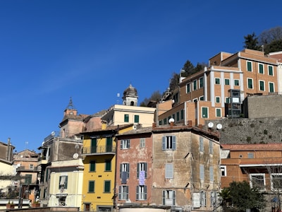A series of colorful, traditional Mediterranean buildings are stacked along a hillside under a clear blue sky. The structures feature a mix of architectural styles, with pastel hues and rustic facades. Windows with shutters, tiled roofs, and satellite dishes are visible, adding character to the scene.
