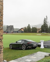 A sleek black sports car parked on a lush green lawn in front of a luxury resort or hotel. The background features buildings with classic architecture, surrounded by tall trees and a misty mountainous landscape. Rain droplets are visible on the car, indicating wet weather. A white tall table with a small object on top is nearby, and a golf course is visible in the distance.