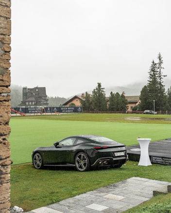 A sleek black sports car parked on a lush green lawn in front of a luxury resort or hotel. The background features buildings with classic architecture, surrounded by tall trees and a misty mountainous landscape. Rain droplets are visible on the car, indicating wet weather. A white tall table with a small object on top is nearby, and a golf course is visible in the distance.
