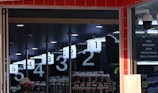 An interior view of a store with checkout lanes numbered from one to five. A man in a hat stands near the entrance. Overhead signage lists categories like toys, stationery, dairy, and frozen meals. Surveillance cameras are visible on the ceiling, and there are merchandise displays in the background.