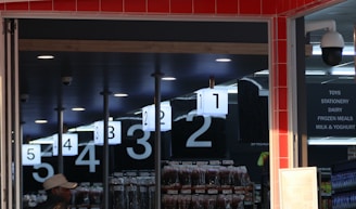 An interior view of a store with checkout lanes numbered from one to five. A man in a hat stands near the entrance. Overhead signage lists categories like toys, stationery, dairy, and frozen meals. Surveillance cameras are visible on the ceiling, and there are merchandise displays in the background.