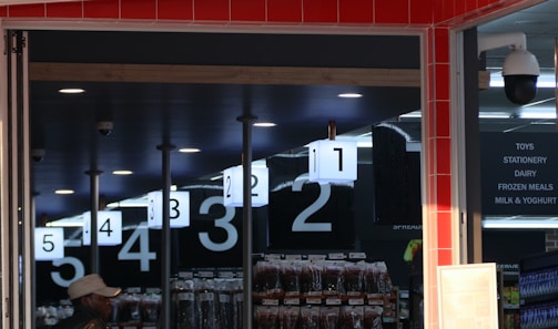 An interior view of a store with checkout lanes numbered from one to five. A man in a hat stands near the entrance. Overhead signage lists categories like toys, stationery, dairy, and frozen meals. Surveillance cameras are visible on the ceiling, and there are merchandise displays in the background.