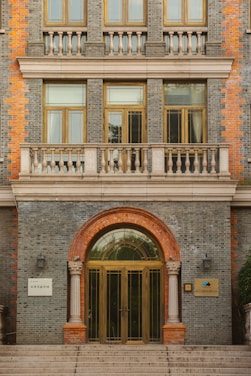 A classical building facade featuring a grand entrance with a semicircular arch surrounded by reddish-brown brickwork. The structure includes intricate columns flanking the entrance and several large windows above, bordered by a balcony with a balustrade. The facade combines gray brick with accents of red bricks.