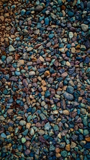 A collection of small, multicolored pebbles tightly packed together, creating a textured and visually rich surface. The stones vary in shape and size, with hues ranging from deep blues and purples to earthy browns and greens.