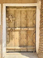 A wooden door with intricate carvings and geometric patterns, featuring a rustic latch and handle. The texture of the wood is aged and weathered, surrounded by a white frame. Both sides of the door reveal the texture of earthen walls.