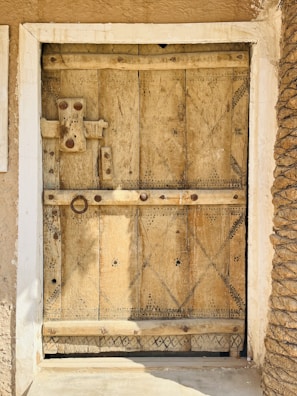 A wooden door with intricate carvings and geometric patterns, featuring a rustic latch and handle. The texture of the wood is aged and weathered, surrounded by a white frame. Both sides of the door reveal the texture of earthen walls.