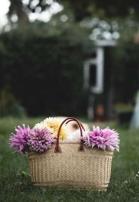 A woven basket with leather handles is filled with large, colorful flowers. The flowers include shades of purple, pink, and yellow, creating a vibrant contrast against the natural texture of the basket. The basket is placed on green grass in a lush garden setting.