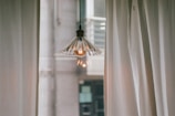 A vintage-style hanging lamp with a clear, glass shade is centered between two white curtains. The soft glow from the bulb casts a warm light, contrasting with the muted tones of the urban background visible through a window.