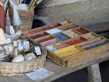 A rustic table holds a wooden tray filled with a variety of colorful pigments in small sections. To the side, there is a wicker basket containing brushes. Several small bottles and jars, some with handwritten labels, are placed on the table, along with a garlic bulb and a small white ball. The setting suggests an artisanal or historical context, possibly for crafting or painting.