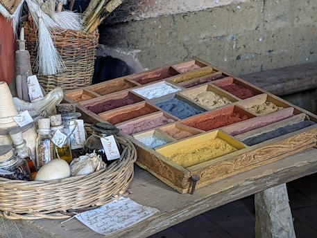 A rustic table holds a wooden tray filled with a variety of colorful pigments in small sections. To the side, there is a wicker basket containing brushes. Several small bottles and jars, some with handwritten labels, are placed on the table, along with a garlic bulb and a small white ball. The setting suggests an artisanal or historical context, possibly for crafting or painting.