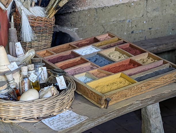 A rustic table holds a wooden tray filled with a variety of colorful pigments in small sections. To the side, there is a wicker basket containing brushes. Several small bottles and jars, some with handwritten labels, are placed on the table, along with a garlic bulb and a small white ball. The setting suggests an artisanal or historical context, possibly for crafting or painting.