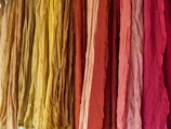 A vibrant display of colorful fabrics hanging vertically, ranging in shades from yellow to deep red. The fabrics appear to have a textured surface with slight wrinkles, suggesting natural fiber material.